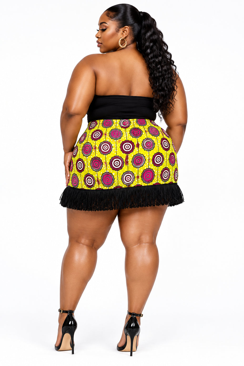 Vista posterior de una minifalda de diseño independiente confeccionada en Ecuador con tela africana Ankara de patrón geométrico circular en tonos amarillo lima, fucsia y negro, terminada con flecos negros en el ruedo inferior.