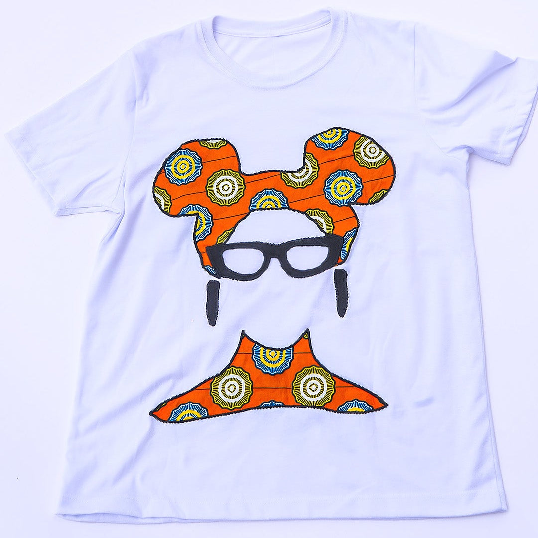 Camiseta blanca de diseño independiente hecha en Ecuador, con una ilustración frontal sutil de un rostro femenino que destaca por su turbante y cuello confeccionados con auténtica tela africana Ankara en patrón geométrico naranja, amarillo y azul, complementada con gafas y aretes negros texturizados.