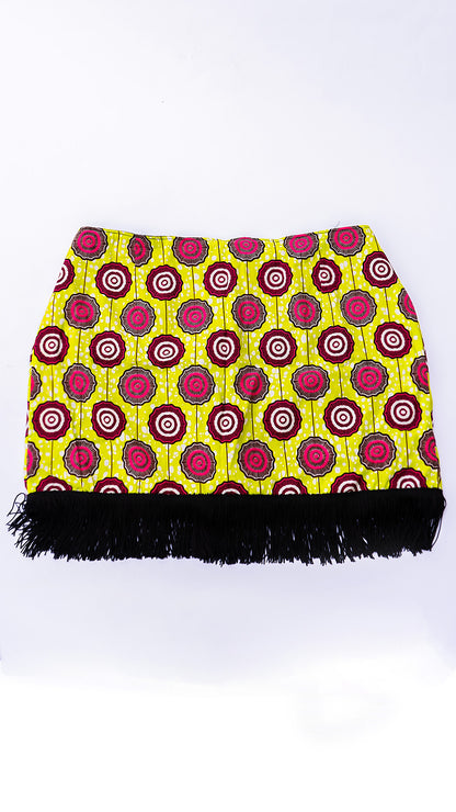 Vista delantera de una minifalda de diseño independiente confeccionada en Ecuador con tela africana Ankara de patrón geométrico circular en tonos amarillo lima, fucsia y negro, terminada con flecos negros en el ruedo inferior.