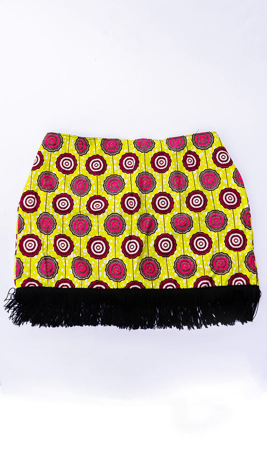 Vista delantera de una minifalda de diseño independiente confeccionada en Ecuador con tela africana Ankara de patrón geométrico circular en tonos amarillo lima, fucsia y negro, terminada con flecos negros en el ruedo inferior.