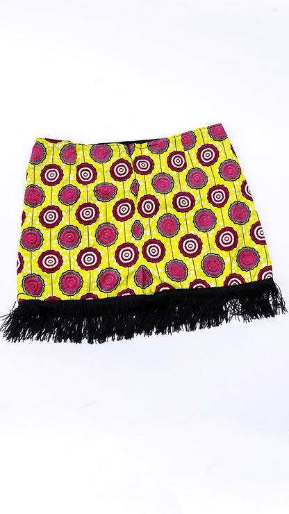Vista posterior de una minifalda de diseño independiente confeccionada en Ecuador con tela africana Ankara de patrón geométrico circular en tonos amarillo lima, fucsia y negro, terminada con flecos negros en el ruedo inferior.